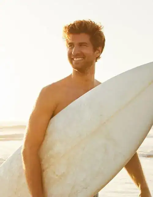 Smiling man holding a surfboard at the beach in warm sunlight.