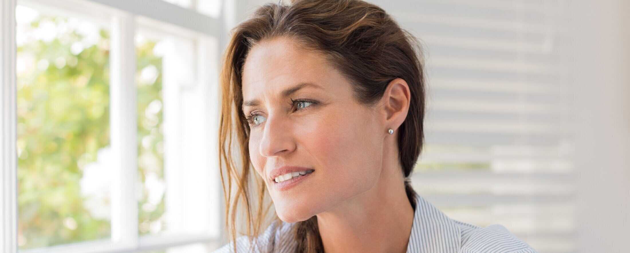 “Woman with healthy, natural-looking skin standing by a window in soft natural light.