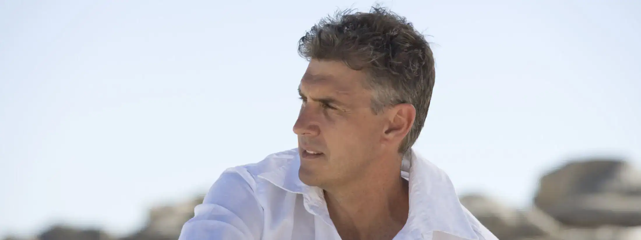 Mature man in a white linen shirt looking to the side with a bright, blurred background.