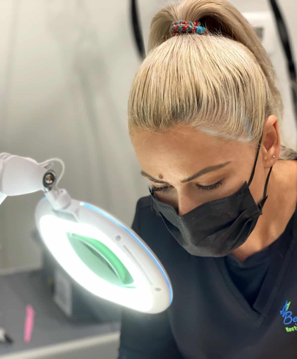 A professional esthetician wearing a black mask and scrubs, using a lighted magnifying lamp to examine a patient's skin.