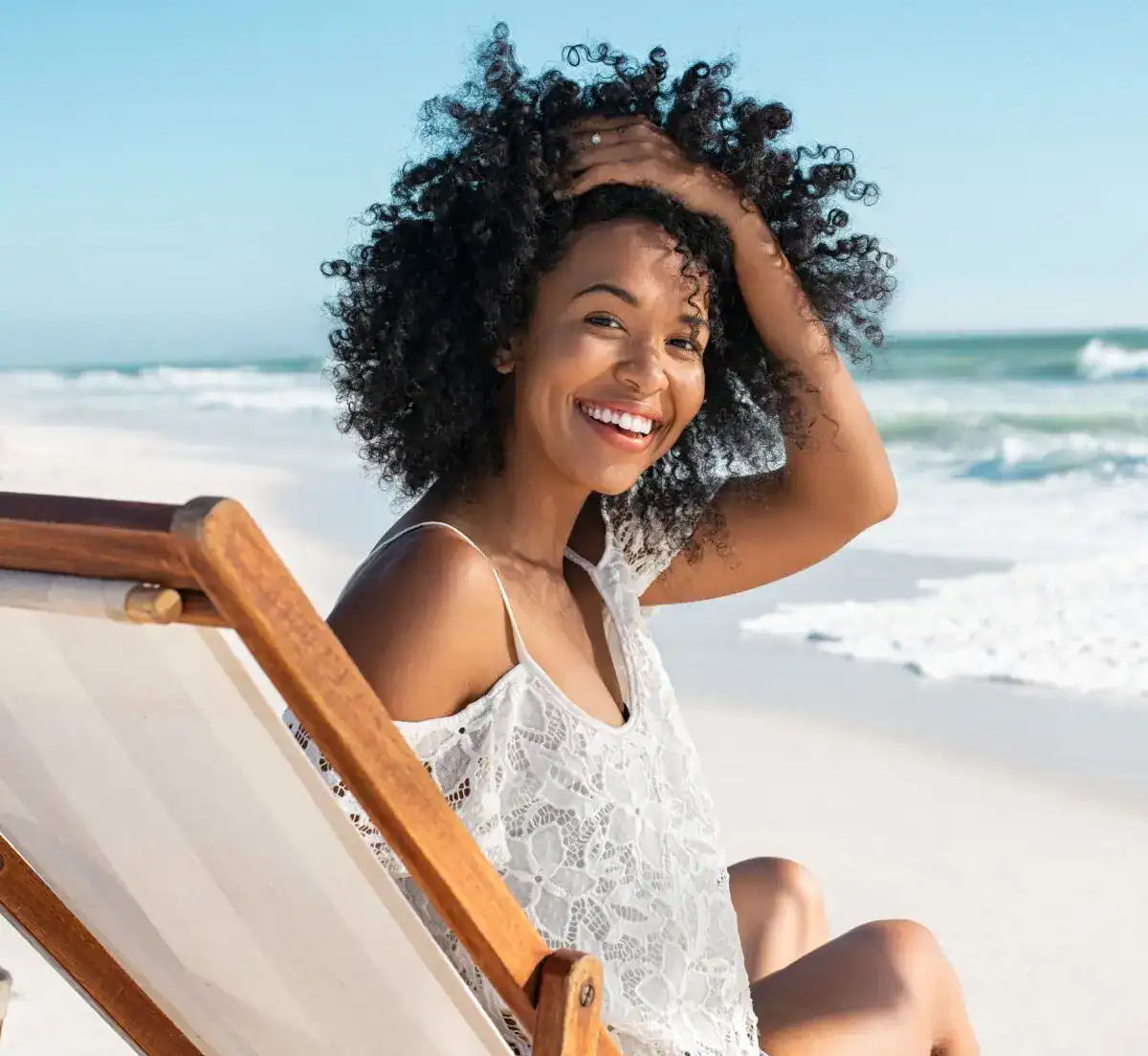 Smiling woman with radiant skin and curly hair relaxing in a beach chair on a white sand shore.