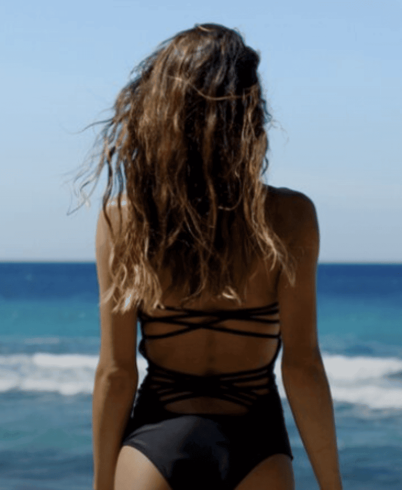 Woman in a black one-piece swimsuit standing by the ocean, viewed from behind