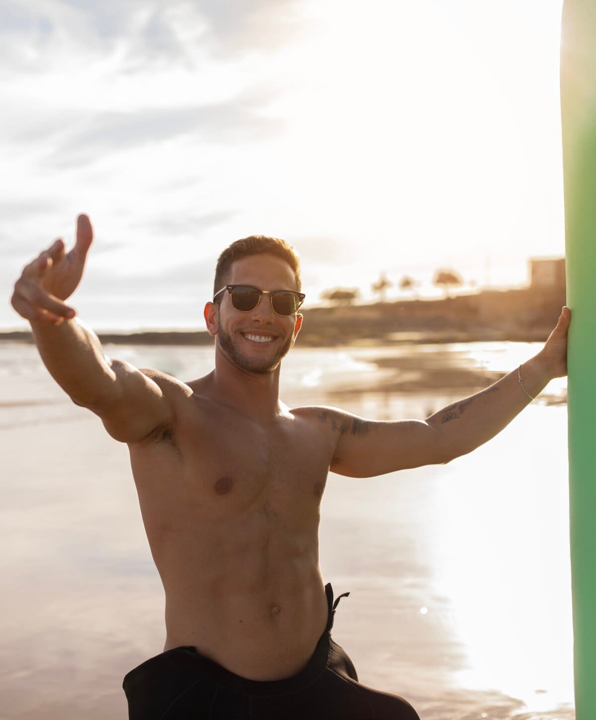 Fit man smiling at the beach holding a surfboard at sunset
