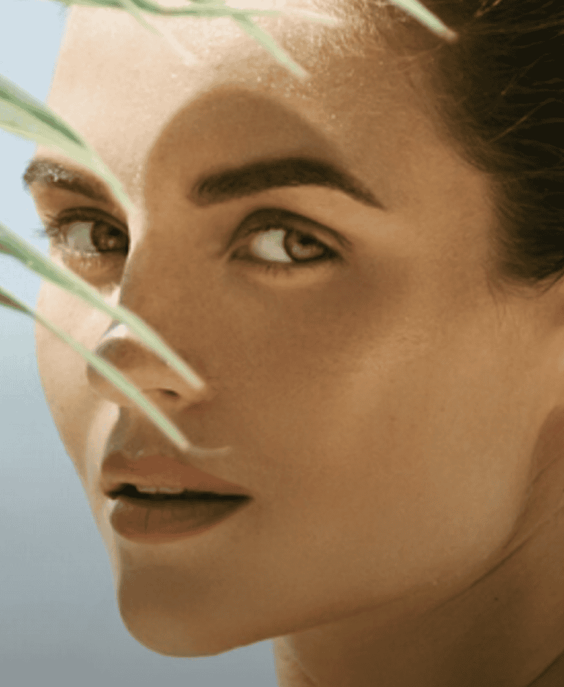 Woman with natural glowing skin posing behind palm leaves on a tropical beach.