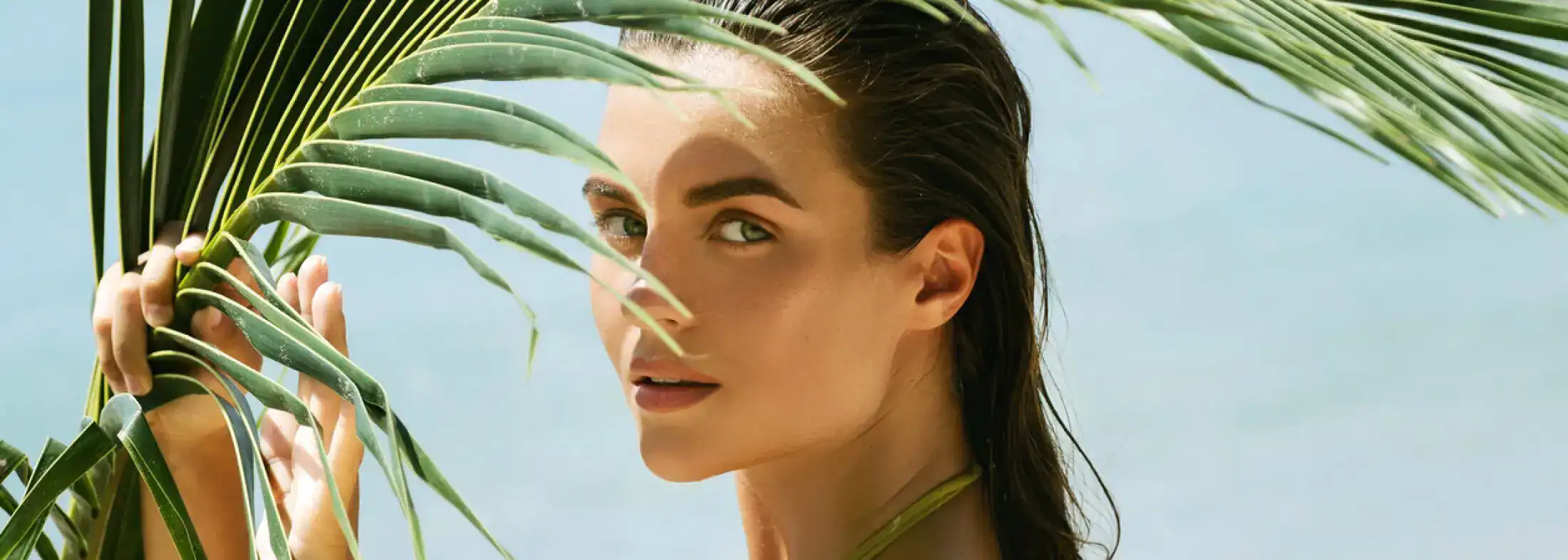 Close-up beauty portrait of a woman with a glowing complexion posing behind tropical palm leaves at the beach.