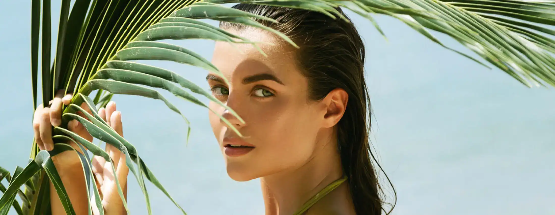 A young woman with wet hair and glowing skin peering through a green palm leaf with a blue ocean background.