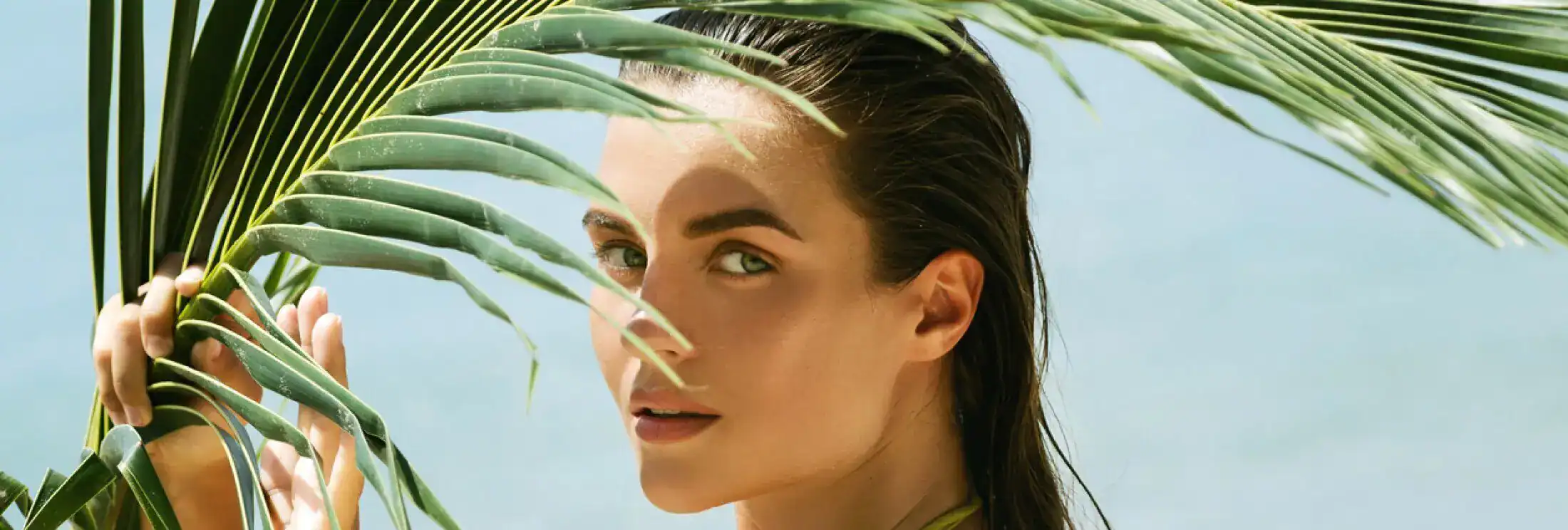 Woman with wet hair posing behind a palm leaf at the beach.