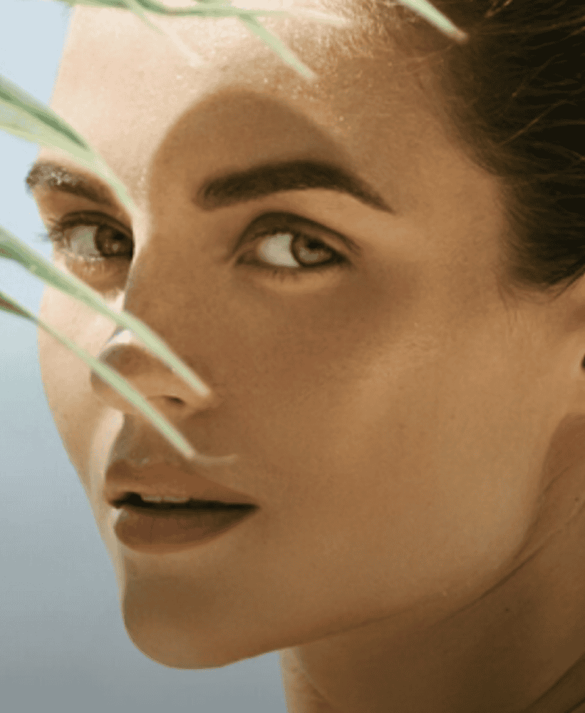 Woman with radiant skin behind palm leaves at the beach.