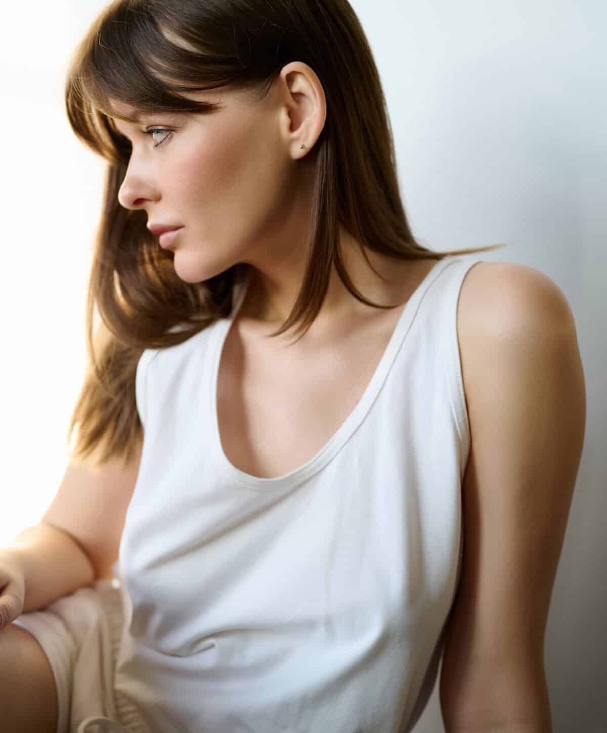 Profile view of a woman with smooth skin and brown hair wearing a white tank top sitting indoors.