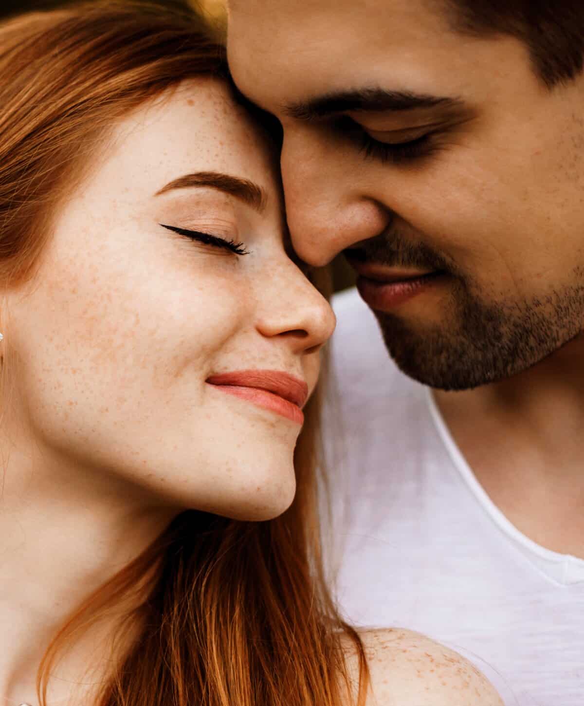 Close-up of a smiling couple with their foreheads touching; the woman has red hair and freckles.