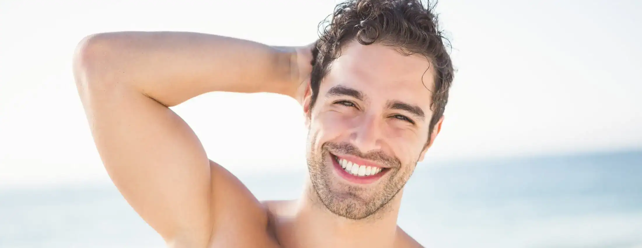Smiling athletic man with curly hair posing on a sunny beach.
