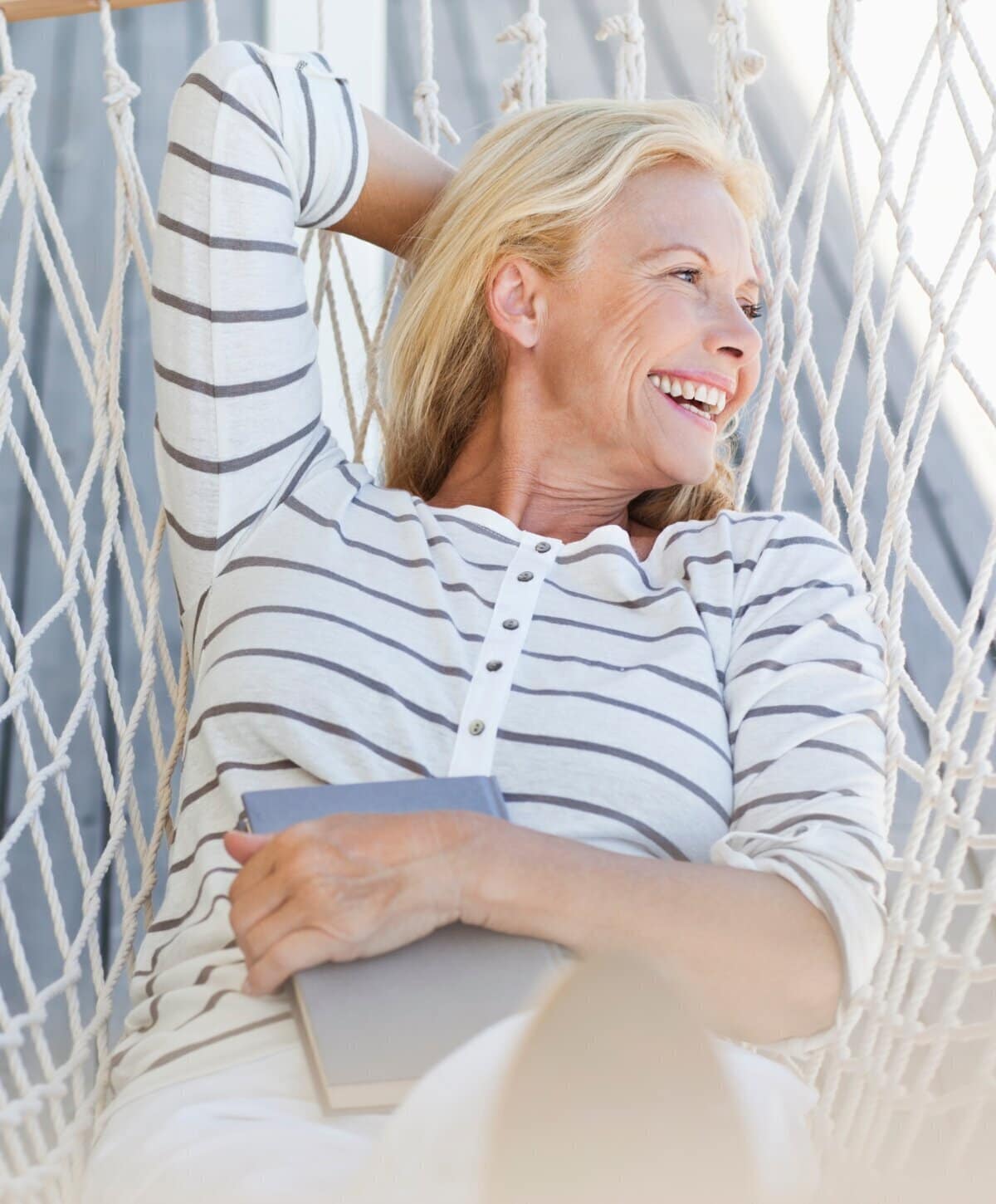 Happy mature blonde woman laughing while relaxing in a rope hammock with a book.