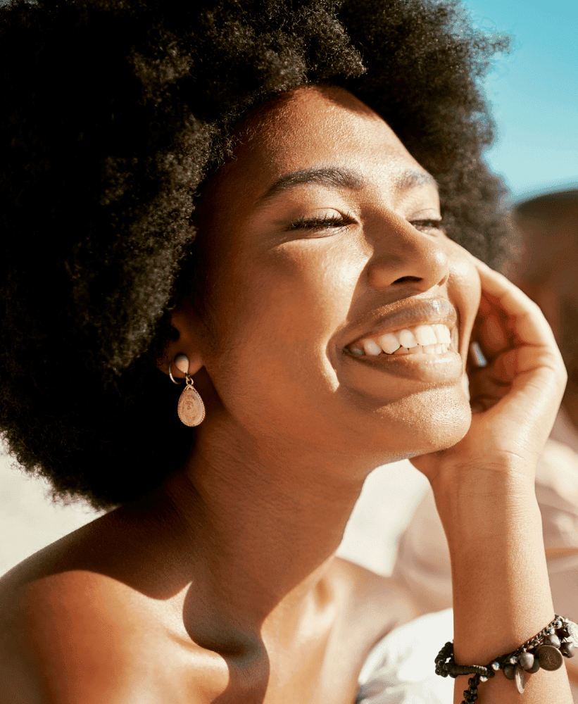 Smiling woman with natural hair enjoying sunlight, representing healthy skin and confidence.