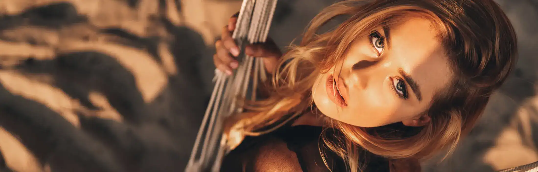 Portrait of a blonde woman with blue eyes lounging in a hammock on a sandy beach during golden hour.