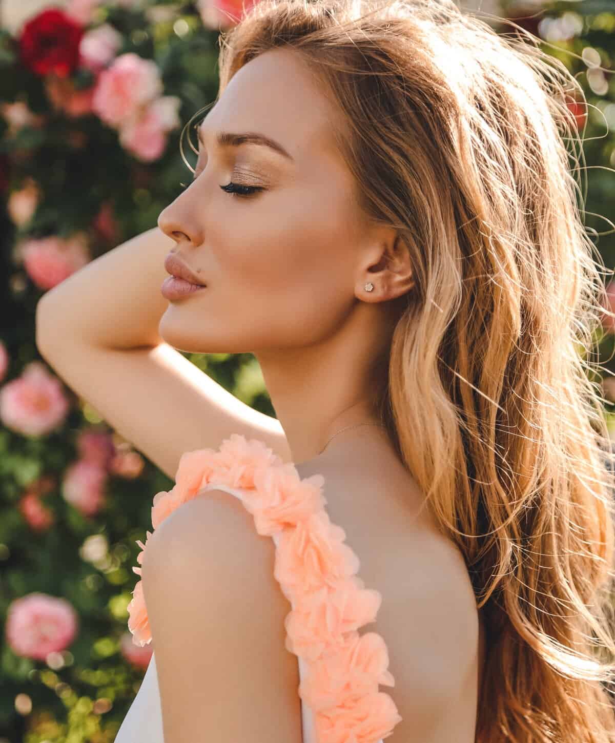 Woman with long blonde hair posing in a sunlit rose garden.
