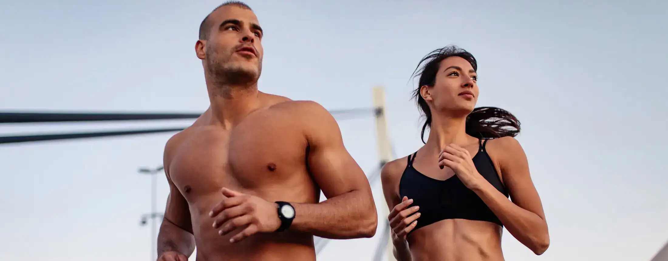 An athletic man and woman jogging outdoors during sunset with a bridge in the background.