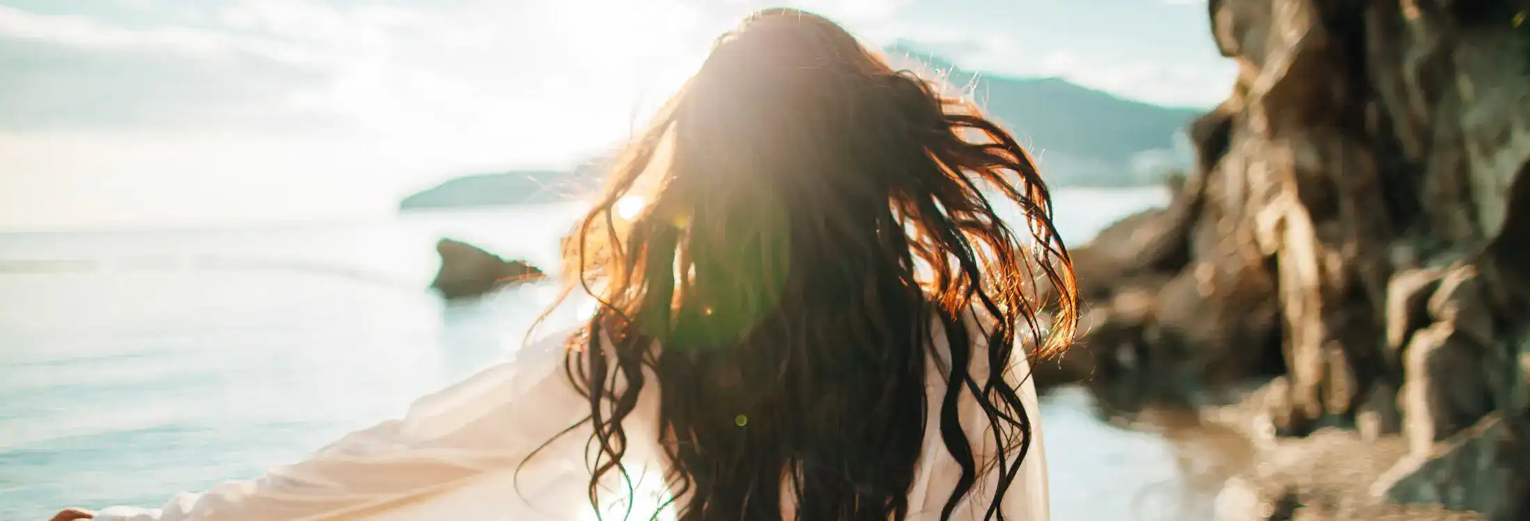Woman with long wavy hair facing a beach sunset symbolizing overall wellness and inner peace.