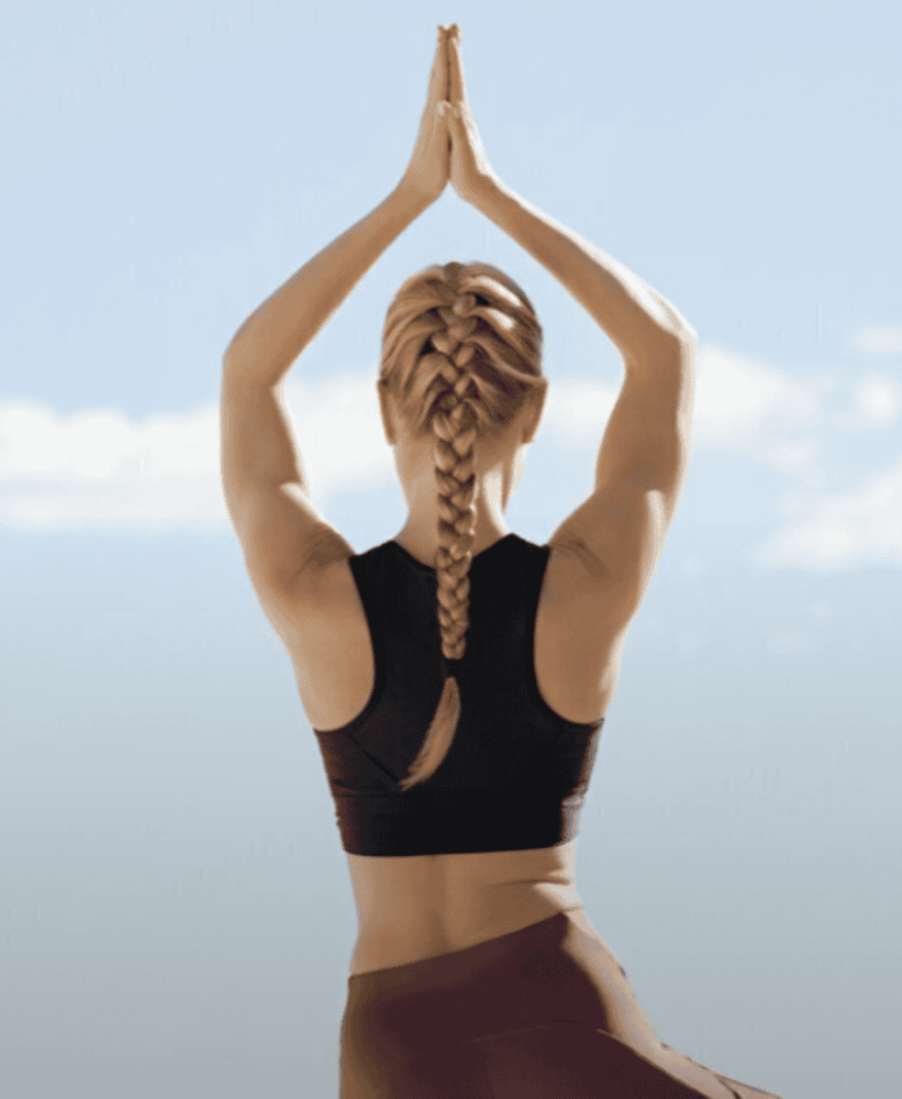 Back view of a woman with a blonde braid performing a yoga tree pose outdoors.