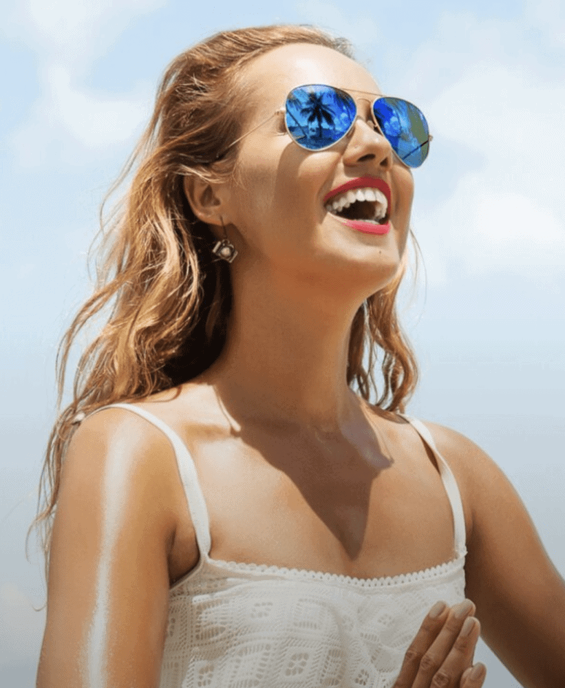 Smiling woman in a white lace top wearing blue mirrored aviator sunglasses reflecting palm trees.